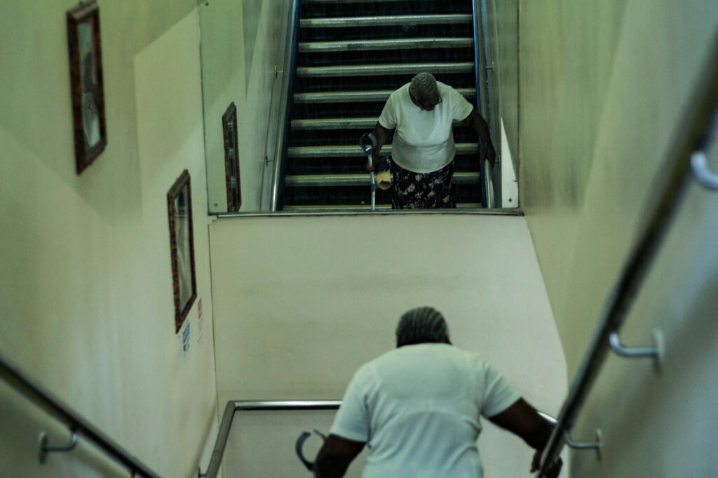 An elderly woman using a cane descends stairs, reflected in a mirror on the wall.