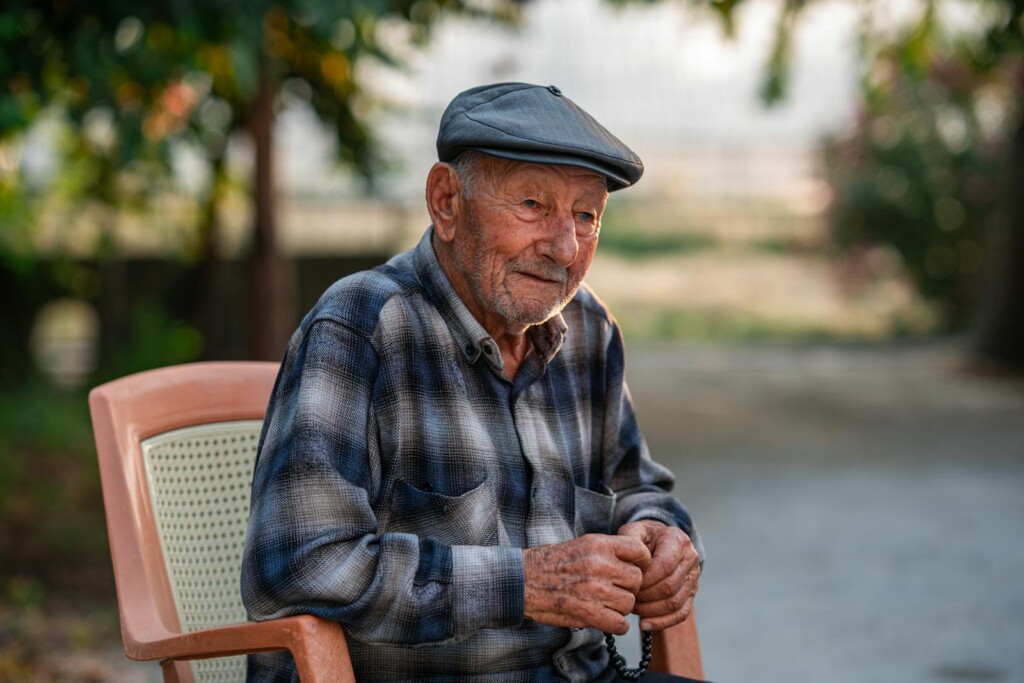 Senior adult man seated outdoors wearing a cap and plaid shirt, exuding tranquility.